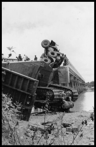 A picture taken at riverside level of A Scammell Constructor Lowloader lies on it's side perched precariously over the river.