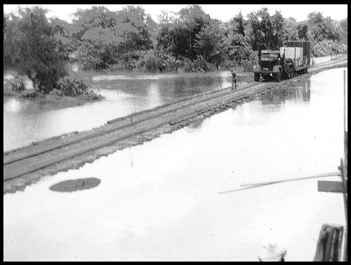 A giant Scammell lowloader crosses a long wooden bridge over flooded rice paddy.